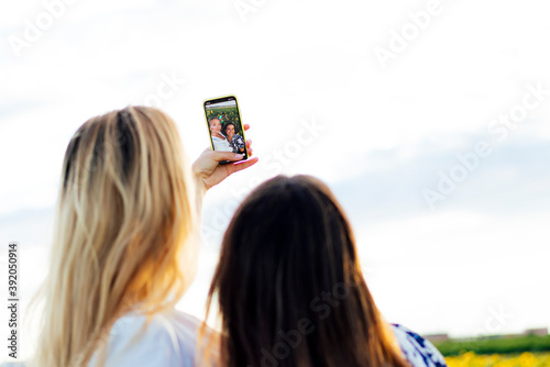 A couple of attractive young women one blonde and the other brunette posing in their designer dresses in a field of sunflowers using their smartphone photo