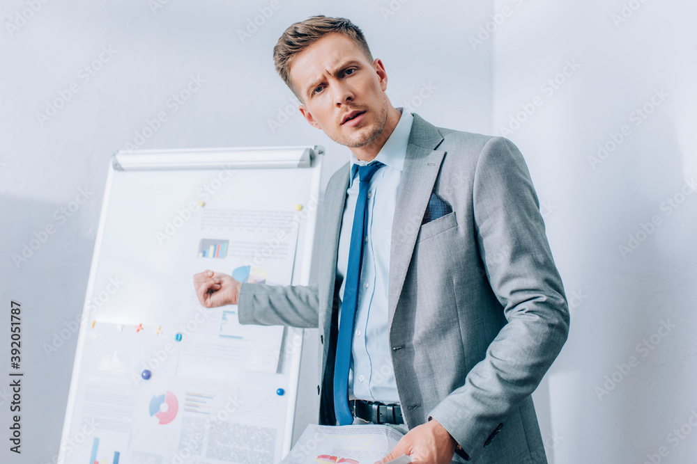 Focused businessman looking at camera while pointing at flipchart on blurred background in office