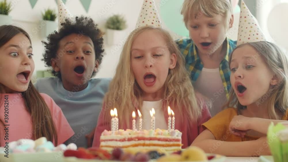 Medium close-up of Caucasian blond-haired girl counting with hand gestures to start blowing out candles on cake with diverse school friends. Children laughing, clapping hands on party indoors