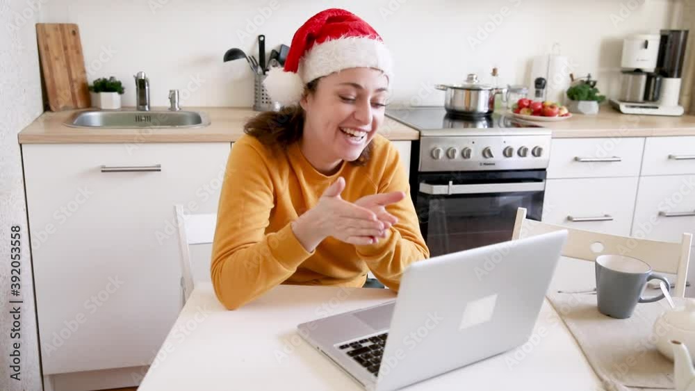 Smiling girl wearing Santa hat waving hand video calling family by webcam. Woman with laptop having virtual meeting chat on holidays sitting on kitchen at home. Happy Christmas and New Year new normal Smiling girl wearing Santa hat waving hand video calling family by webcam. Woman with laptop having virtual meeting chat on holidays sitting on kitchen at home. Happy Christmas and New Year new normal