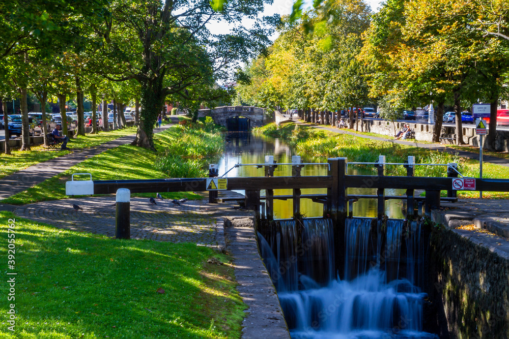 Fototapeta premium Grand Canal in Dublin on sunny summer day with people sitting on benches enjoying sunshine. Water flows over lock gates at Conyngham Bridge from Huband Bridge. Ireland