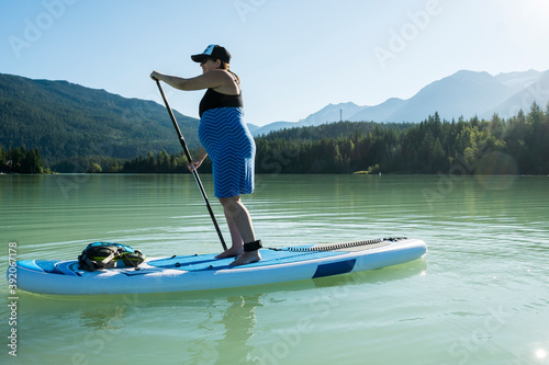 Full body pregnant woman in dress paddleboarding on calm water of lake against mountain ridge and green forest on summer day in British Columbia, Canada
