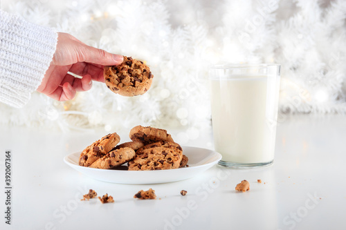 Person holds a homemade chocolate chip cookie accompanied by a glass of milk and illuminated by the lights of a white Christmas tree on a Christmas day