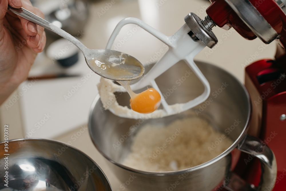 A close photo of the yolk is falling from the spoon into the stainless steel bowl of the red stand mixer with shortcrust pastry. A close photo of the yolk is falling from the spoon into the stainless steel bowl of the red stand mixer with shortcrust pastry.