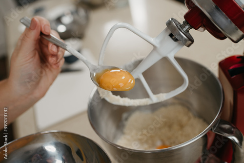 A close photo of the hand of the woman who is holding the spoon with the yolk above the stainless steel bowl of the red stand mixer with shortcrust pastry.