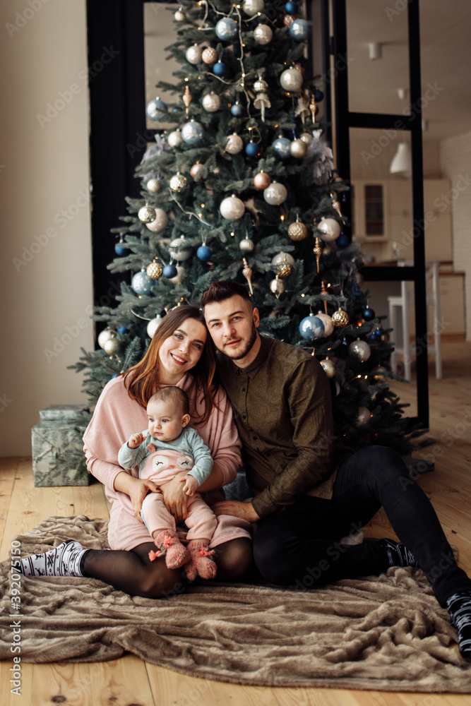 Lovely parents with cute kid sit near Christmas tree at home, smile, caring mom hold little daughter in arms, family enjoy winter holidays together, New Year celebration concept