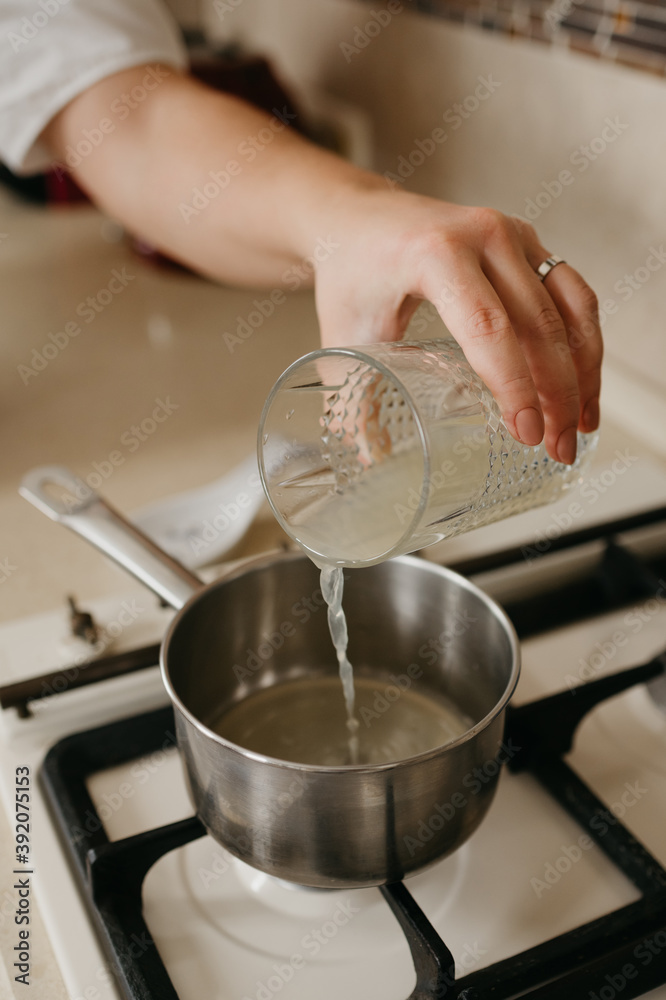 A hand of a woman who is pouring fresh lemon juice from the glass to the saucepan on the gas stove