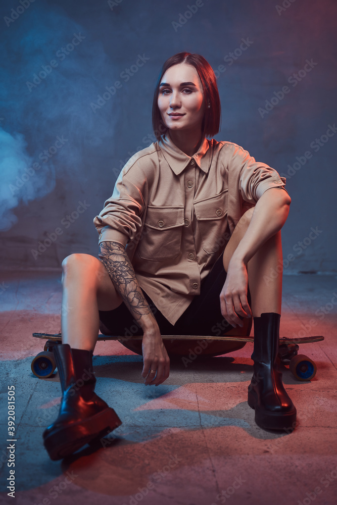 Smiley and cheerful girl with short haircut and dressed in stylish clothing poses sitting on skateboard in smokey background.