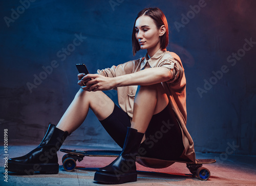Beautiful hipster woman dressed in stylish casual clothing with short brown hairs sits on skate messaging using her smartphone.