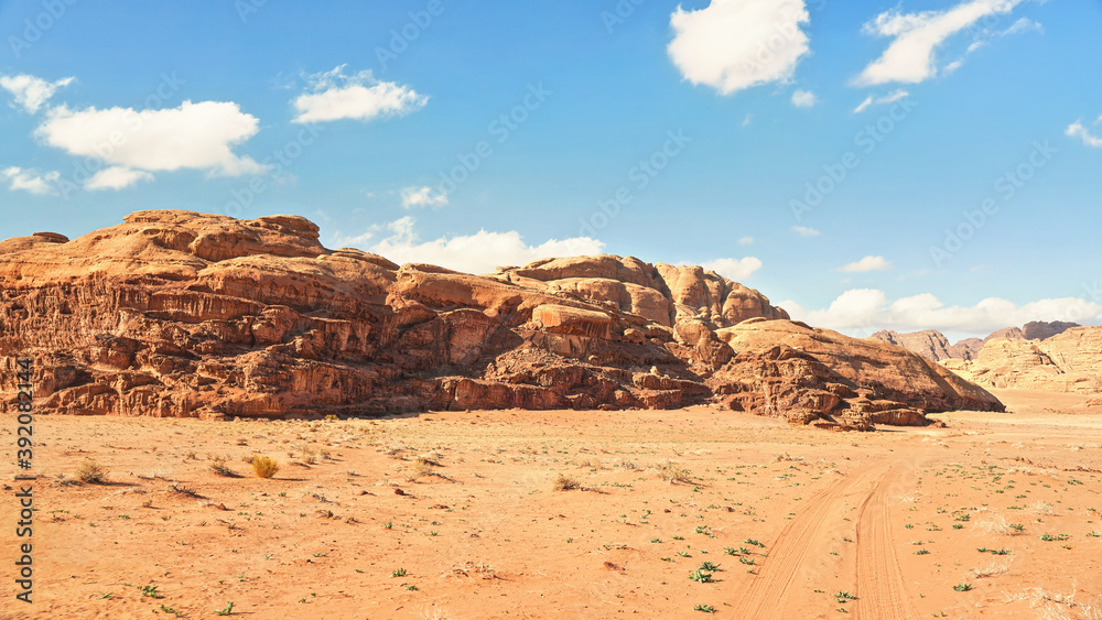 Rocky massifs on red sand desert, vehicle tracks ground, bright cloudy sky in background, typical scenery in Wadi Rum, Jordan Rocky massifs on red sand desert, vehicle tracks ground, bright cloudy sky in background, typical scenery in Wadi Rum, Jordan