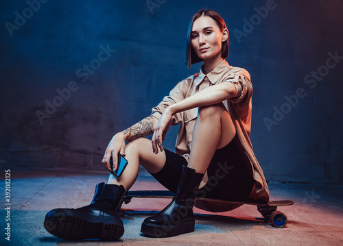 Attractive and stylish woman with tattoo and short haircut poses sitting on skateboard and holding her smartphone.