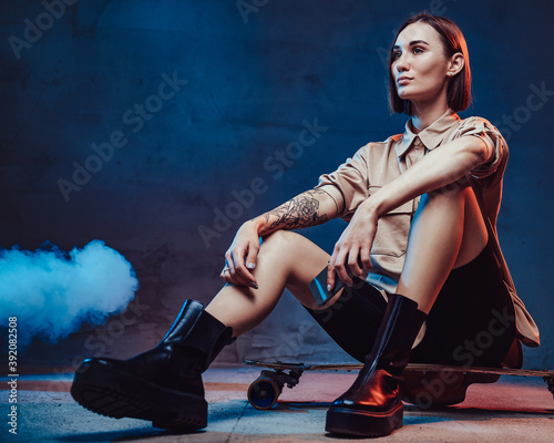Dressed in stylish casual clothing girl with modern smartphone and tattoo on her right hand sits on skate in smokey background.