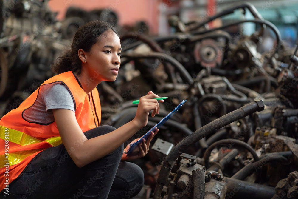 African American female worker in safety vest working with clipboard and checking old automotive spare parts, engine, machine for repair or maintenance in automotive spare parts storage warehouse African American female worker in safety vest working with clipboard and checking old automotive spare parts, engine, machine for repair or maintenance in automotive spare parts storage warehouse