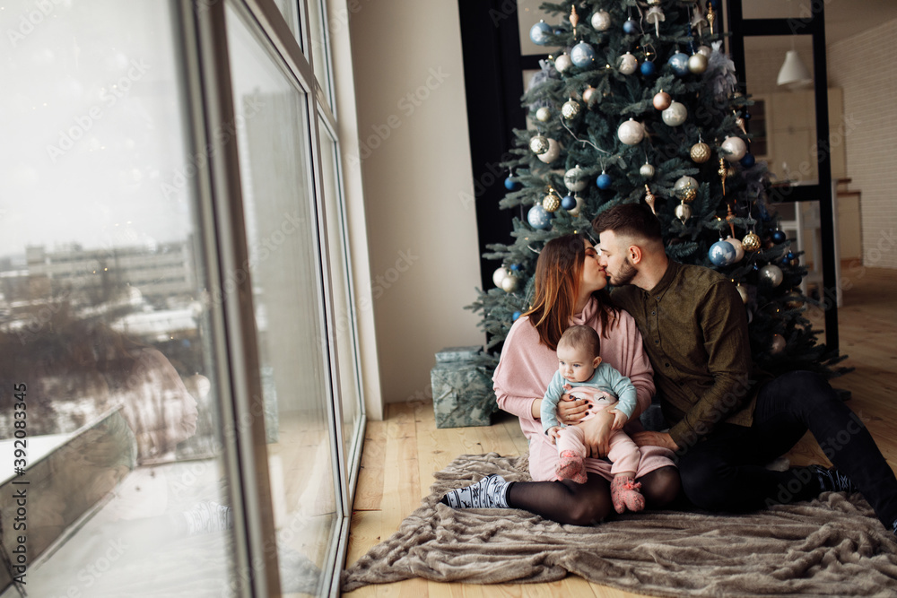 Lovely family sit near the Christmas tree at home, caring mom hold little daughter in arms, loving husband kiss beautiful wife, enjoy winter holidays, New Year celebration concept Lovely family sit near the Christmas tree at home, caring mom hold little daughter in arms, loving husband kiss beautiful wife, enjoy winter holidays, New Year celebration concept