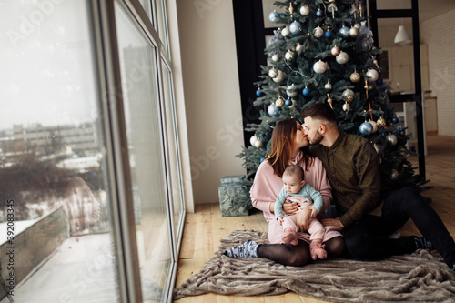 Lovely family sit near the Christmas tree at home, caring mom hold little daughter in arms, loving husband kiss beautiful wife, enjoy winter holidays, New Year celebration concept