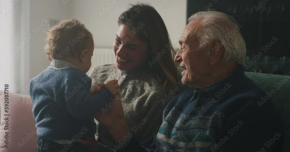 Cinematic shot of happy family: grandfather, daughter and grandson baby are having fun to pass time together while sitting on sofa at home. Concept of life, grandparents, generation, childhood, love