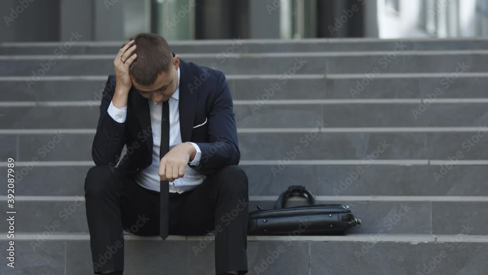 Sad young businessman sitting on the stairs near office building. Tired putting hands to the head. Male office worker in despair lost job. Feeling bad and annoyed.