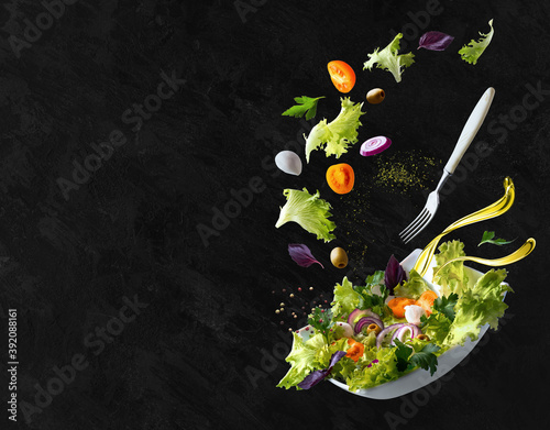 A white plate with salad and floating in the air ingredients: olives, lettuce, onion, tomato, mozzarella cheese, parsley, basil and olive oil. Black background. Copy space.