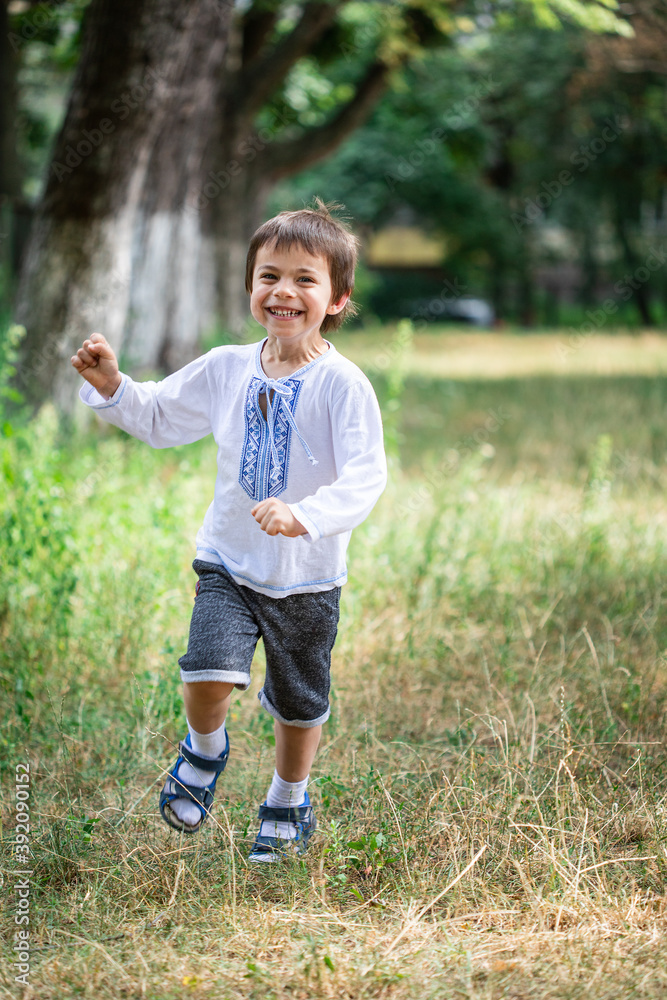 Little adorable boy in shirt with Ukrainian ornament running in park and smiling Little adorable boy in shirt with Ukrainian ornament running in park and smiling