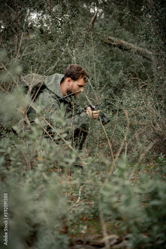 young mountaineer man dressed in dark green in the undergrowth crouching taking photos of the plants, dark and desaturated style, adventure concept.