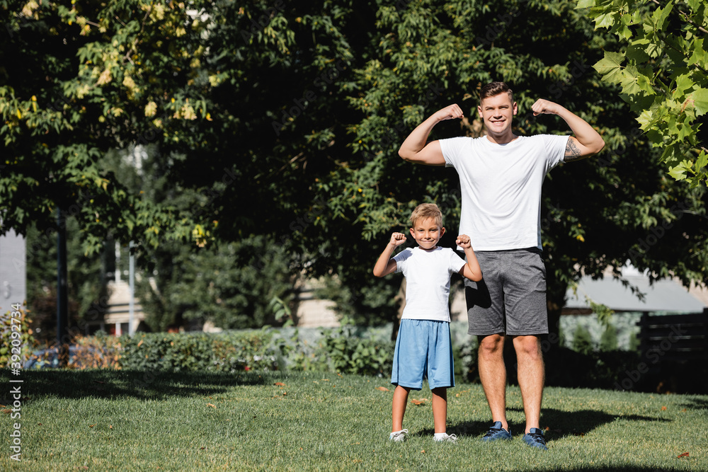 Smiling father and son looking at camera while posing with raised hands showing power in park
