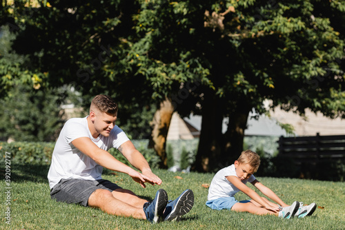 Father and son in sportswear with outstretched hands warming up, while sitting on grass on blurred background