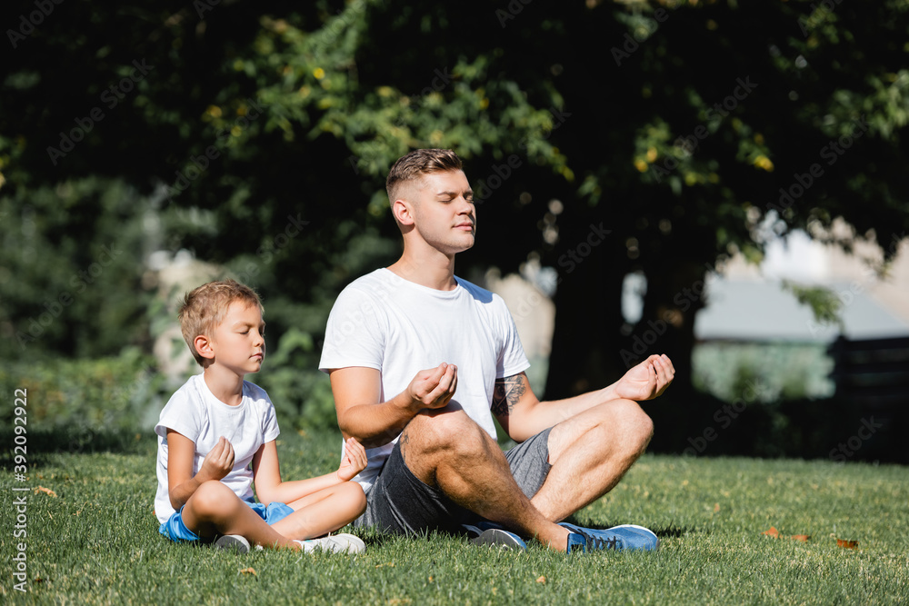 Father and son with closed eyes relaxing while sitting in lotus pose in park on blurred background Father and son with closed eyes relaxing while sitting in lotus pose in park on blurred background