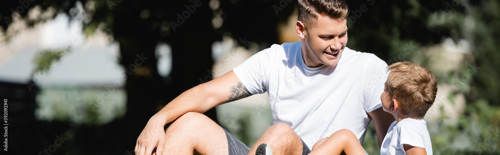 Smiling father sitting and looking at son with blurred trees on background, banner Smiling father sitting and looking at son with blurred trees on background, banner