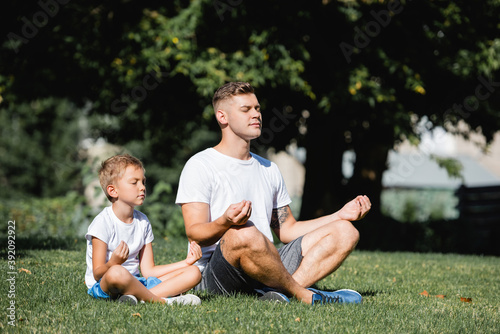 Father and son with closed eyes relaxing while sitting in lotus pose in park on blurred background