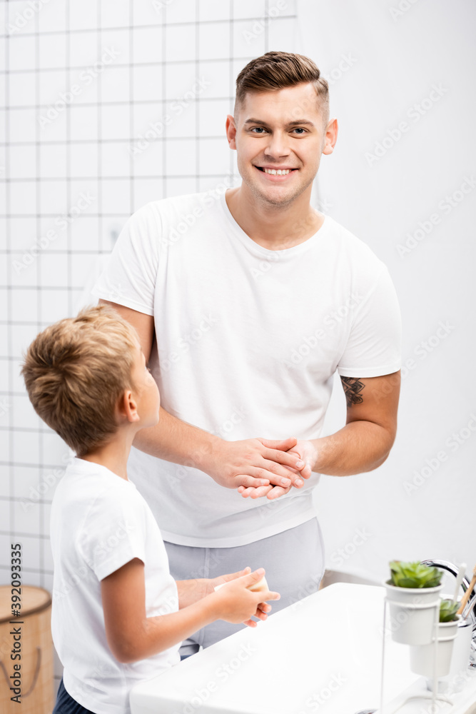 Fototapeta premium Son holding soap and standing near smiling father rubbing hands while looking at camera in bathroom