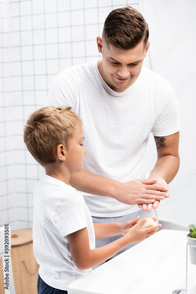 Fototapeta premium Smiling father washing hands, while looking at son with soap standing near sink in bathroom