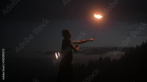 Graceful male fireshow performer making fire dance with two metal fans lit with flames on night mountains meadow