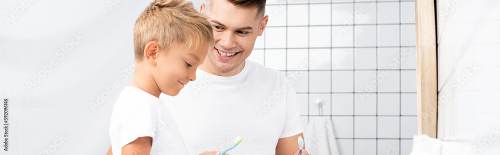 Smiling father looking at son holding toothbrush in bathroom, banner Smiling father looking at son holding toothbrush in bathroom, banner