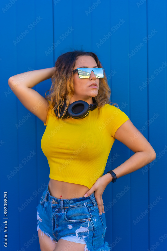 Lifestyle, blonde Caucasian girl with yellow t-shirt on a blue background. Young woman posing with sensual gaze with music headphones and sunglasses