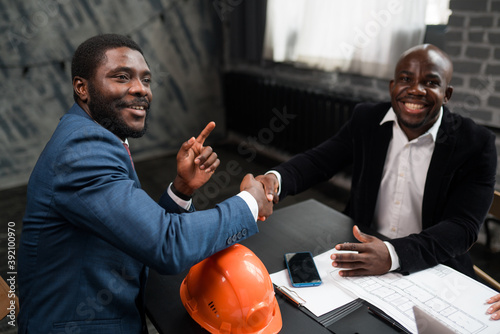 Two African Americans signed a cooperation agreement. Business investors shake hands. Orange construction hard hat on office desk