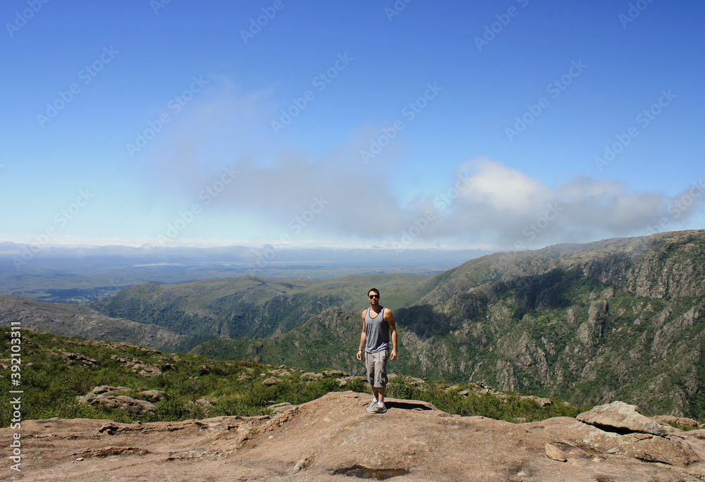 Hiker in the rocky mountaintop. Male young adult trekking the mountain summit. Beautiful view of the rocky cliffs and forest under a blue sky in a sunny summer day. 