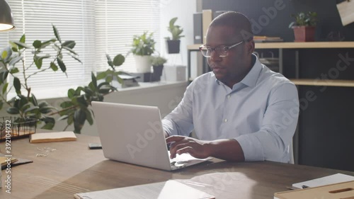 Arc shot of professional African American businessman typing on laptop during workday while sitting at desk in office photo
