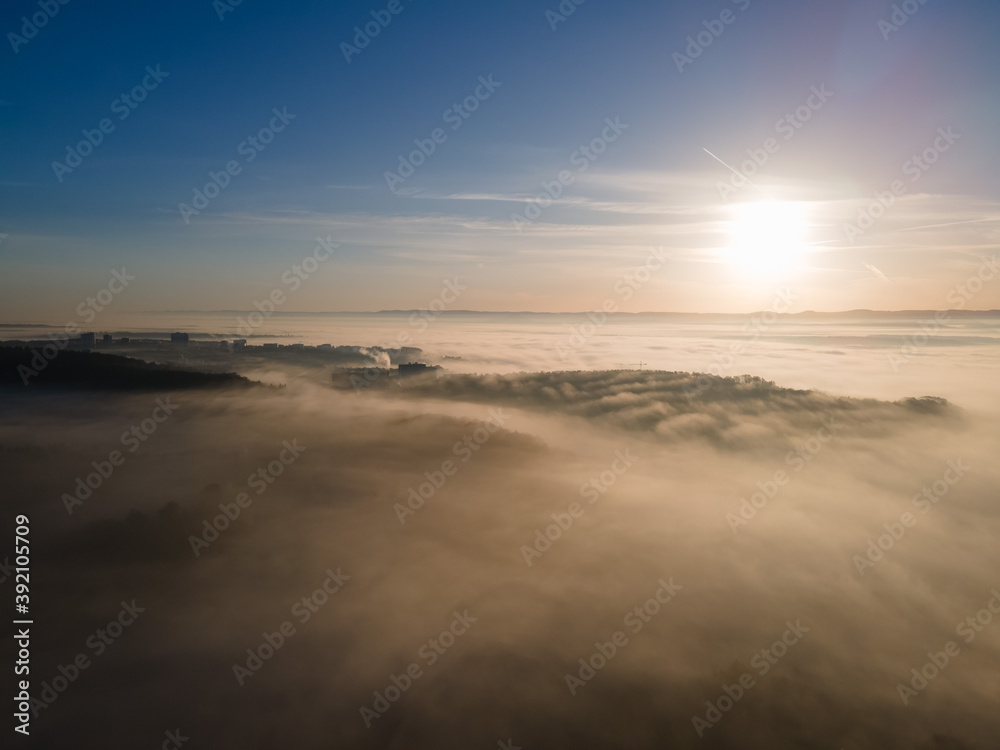 Aerial view of sunrise above foggy town. Mystical fog scattering sunrays of upcoming sun.