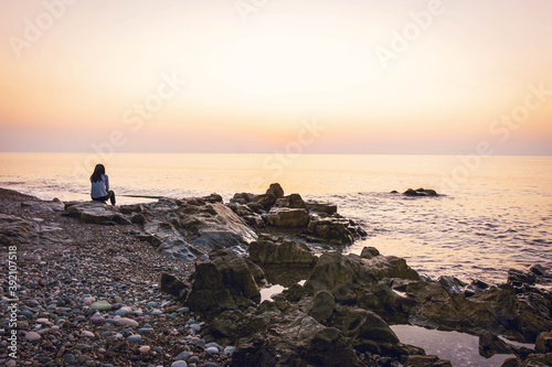 Woman sits on the rock the beach looking to horizon after sunset with calm sea in the background. Solitude and tranquility.