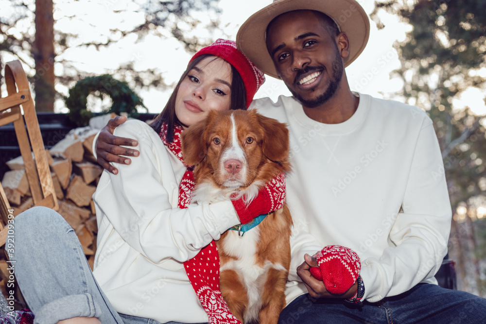 Multiracial couple are sitting in the trunk of a car with a dog. A couple of lovers came to the forest for a Christmas tree.