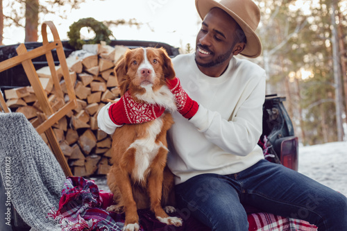 African American man sits with a dog in the trunk of a car in the winter forest. A man in a white sweatshirt.