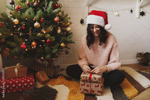 Happy young woman in santa hat wrapping gift under christmas tree in festive decorated room