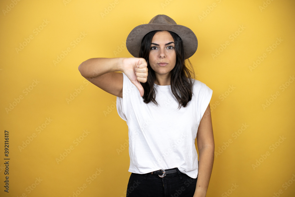 Beautiful woman wearing casual white t-shirt and a hat standing over yellow background with angry face, negative sign showing dislike with thumb down Beautiful woman wearing casual white t-shirt and a hat standing over yellow background with angry face, negative sign showing dislike with thumb down