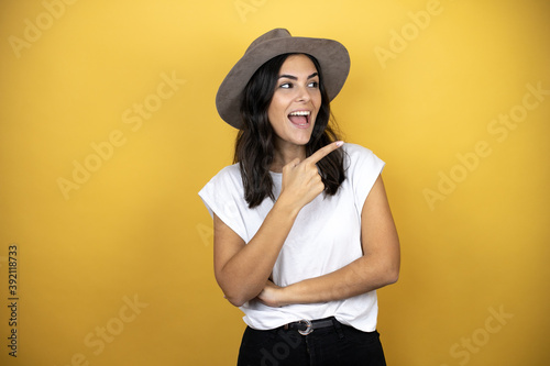 Beautiful woman wearing casual white t-shirt and a hat standing over yellow background smiling and pointing with hand and finger to the side