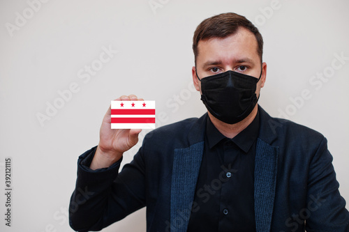 Man wear black formal and protect face mask, hold District of Columbia flag card isolated on white background. USA coronavirus Covid country concept.