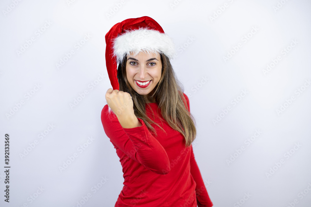 Young beautiful woman wearing a Santa hat over white background angry and mad raising fist frustrated and furious while shouting with anger. Rage and aggressive concept. Young beautiful woman wearing a Santa hat over white background angry and mad raising fist frustrated and furious while shouting with anger. Rage and aggressive concept.