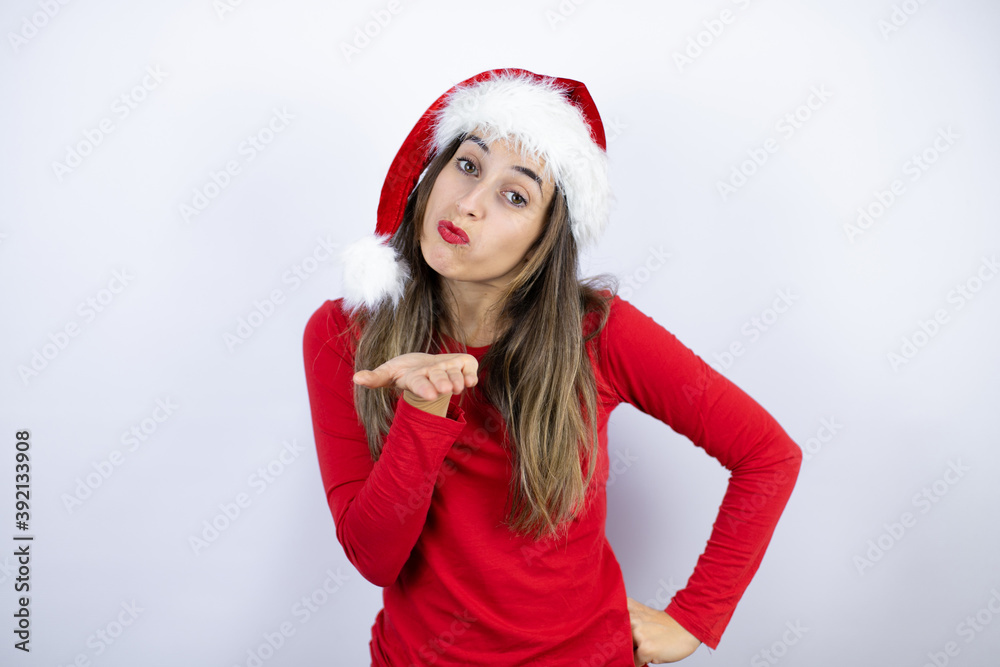 Young beautiful woman wearing a Santa hat over white background looking at the camera blowing a kiss with hand on air being lovely and sexy. Love expression. Young beautiful woman wearing a Santa hat over white background looking at the camera blowing a kiss with hand on air being lovely and sexy. Love expression.