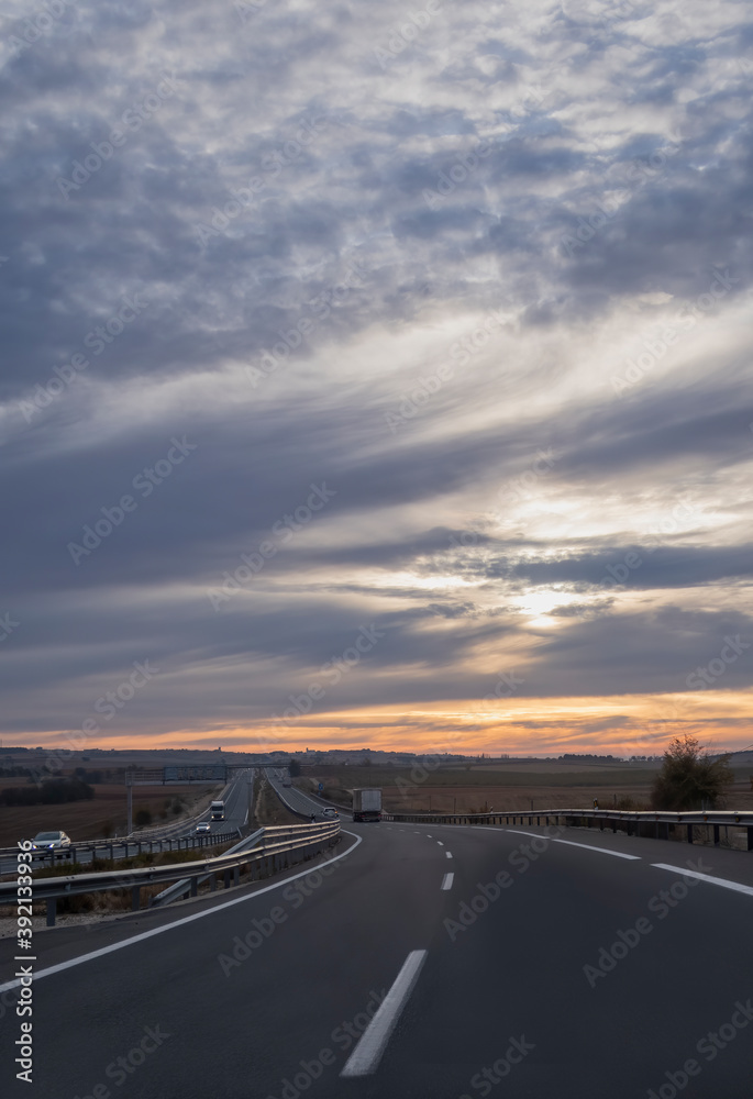 view of a curve in a highway at a cloudy sunrise with a truck in the distance and a village in the background, portrait format
