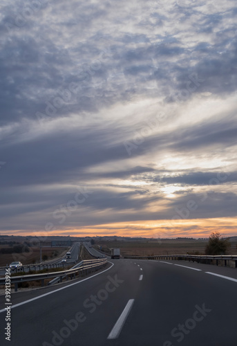 view of a curve in a highway at a cloudy sunrise with a truck in the distance and a village in the background, portrait format