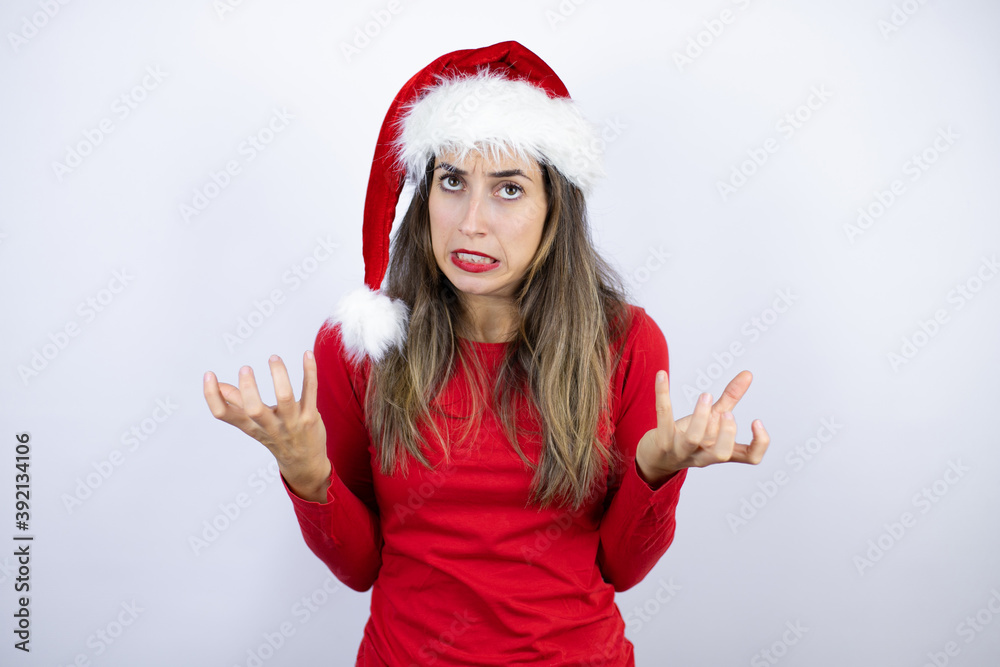 Young beautiful woman wearing a Santa hat over white background crazy and mad shouting and yelling with aggressive expression and arms raised. Frustration concept. Young beautiful woman wearing a Santa hat over white background crazy and mad shouting and yelling with aggressive expression and arms raised. Frustration concept.